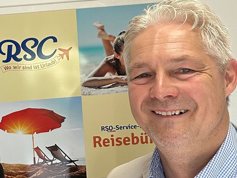 Ein lächelnder Mann mit grauen Haaren im hellen Sakko und kariertem Hemd steht vor einem Poster mit Urlaubs-Motiven. Auf dem Poster sind ein Sonnenschirm, Strandliegen, eine Frau am Strand sowie das Logo „RSC – Wo wir sind ist Urlaub!“ und der Schriftzug „Reisebüro“ zu sehen.