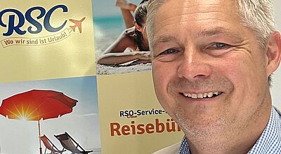 Ein lächelnder Mann mit grauen Haaren im hellen Sakko und kariertem Hemd steht vor einem Poster mit Urlaubs-Motiven. Auf dem Poster sind ein Sonnenschirm, Strandliegen, eine Frau am Strand sowie das Logo „RSC – Wo wir sind ist Urlaub!“ und der Schriftzug „Reisebüro“ zu sehen.