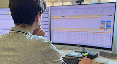 Ein junger Mann sitzt am Schreibtisch vor zwei Computerbildschirmen, auf denen Tabellen und Informationen angezeigt werden. Er trägt ein graues Polohemd und stützt sein Kinn nachdenklich auf eine Hand, während er mit der anderen Hand die Computermaus bedient. Im Hintergrund fällt Tageslicht durch das Fenster auf den Arbeitsplatz, auf dem Tastatur, Taschenrechner und weitere Büroutensilien liegen.