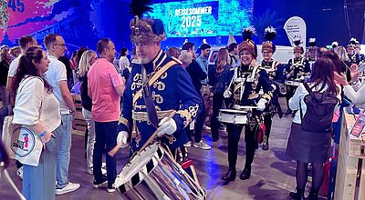 Trommelwirbel zum Auftakt: Der zweite QTA-Reisesommer war mit Abstand das größte Vertriebs-Event des Jahres. Eine Gruppe Trommler in Garde-Uniformen laufen durch eine Messehalle, Besucher daneben.