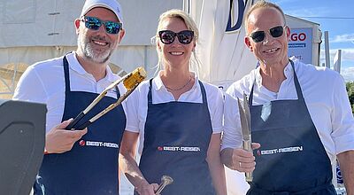 Zwei Männer und eine Frau mit blauen Kochschürzen und Grillzangen in der Hand lachen in die Kamera. Sie stehen neben einem Grill, die Sonne scheint. Auf den Schürzen steht Best-Reisen