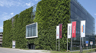Modernes Bürogebäude mit Glasfassade, dessen eine Seite komplett von einer grünen Pflanzenwand bedeckt ist; davor wehen mehrere Fahnen mit den Logos von Hotelplan, Migros und anderen Marken.