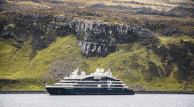 Ein Yacht-ähnliches Luxuskreuzfahrtschiff mit dunkelblauem Bug und weißem, schnittigen Aufbau liegt vor einer britischen Insel auf Reede. Die Kulisse bilden schroffe Felsen in verschiedenen Grautönen und karge grüne Wiesen