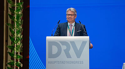 Ein Manager mittleren Alters in einem grauen Anzug steht lächelnd auf einem Rednerpult mit der Aufschrift DRV vor einer blauen Leinwand