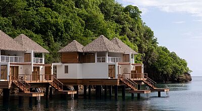 Zweistöckige Overwater-Villen mit Reetdach vor tropisch-bewaldetem Berghang