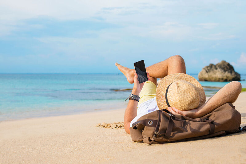 Ein Urlauber mit Strohhut liegt bei Sonnenschein an einem Strand und schaut auf sein Smartphone.