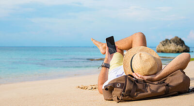 Die neue Ziel-App hilft Reisebüro-Kunden vor und während des Urlaubs. Ein Urlauber mit Strohhut liegt bei Sonnenschein an einem Strand und schaut auf sein Smartphone.
