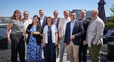 Eine Gruppe von zehn Personen steht bei sonnigem Wetter auf einer Dachterrasse vor städtischer Kulisse. Die Menschen sind unterschiedlich gekleidet – von Business-Outfits bis zu sommerlicher Freizeitkleidung – und lächeln in die Kamera. Im Hintergrund sind moderne Gebäude und Sonnenschirme zu sehen. Das Bild wirkt freundlich und professionell.