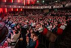 Blick in ein dunkles Theater mit sehr vielen Menschen, die auf eine Bühne schauen