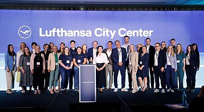 Eine große Gruppe von etwa 30 Personen steht auf einer Bühne vor einer blauen Wand mit dem Logo und Schriftzug „Lufthansa City Center“. Die Personen sind überwiegend geschäftlich gekleidet, einige tragen Firmenkleidung mit Namensschildern und Lanyards. In der Mitte steht ein Rednerpult mit dem Lufthansa City Center Logo. Die Stimmung wirkt professionell und zugleich kollegial. Die Veranstaltung findet in einem modernen Konferenzraum statt.