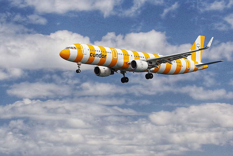 Passagierflugzeug der Fluggesellschaft Condor im gelb-weiß gestreiften Design fliegt über das Meer vor blauem Himmel mit vereinzelten Wolken.
