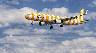 Passagierflugzeug der Fluggesellschaft Condor im gelb-weiß gestreiften Design fliegt über das Meer vor blauem Himmel mit vereinzelten Wolken.