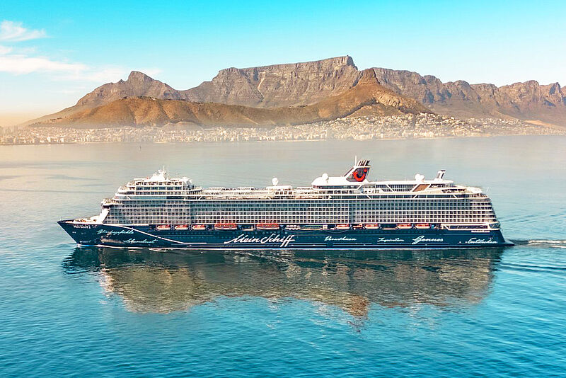 Ein Kreuzfahrtschiff mit dunkelblauem Rumpf fährt bei schönem Wetter vor der Kulisse Kapstadts. Unter blauem Himmel thront der Tafelberg