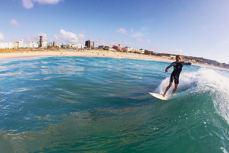 Bondi Beach bei Sydney: Urlauber profitieren vom guten Wechselkurs. Ein Surfer reitet auf einer Welle vor Bondi Beach bei Sydney, Australien, bei sonnigem Wetter; im Hintergrund sind der Sandstrand, Badende und die Skyline mit Gebäuden sichtbar.