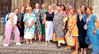 Gruppenfoto von rund 16 Frauen vor einem historischen Gebäude mit schmiedeeisernem Tor. Die Teilnehmerinnen tragen farbenfrohe Sommerkleidung und lächeln in die Kamera.