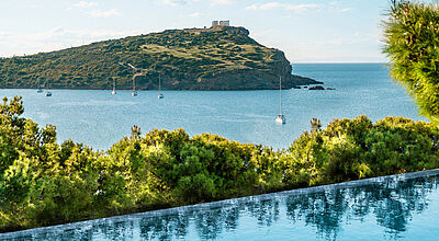 Infinity-Pool mit Blick auf das Meer, mehrere Segelboote und eine grüne Halbinsel mit dem antiken Tempel des Poseidon auf der Spitze, unter blauem Himmel.