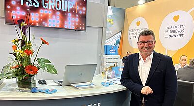 Messestand TSS Group: Mann vor Empfangstresen, Bildschirm mit Firmenlogo, Plakat "Wir lieben Reisebüros"