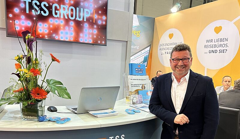 Messestand TSS Group: Mann vor Empfangstresen, Bildschirm mit Firmenlogo, Plakat "Wir lieben Reisebüros"