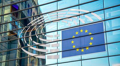 Fassade des Europäischen Parlaments in Brüssel mit dem Emblem des Parlaments und der EU-Flagge – zu sehen sind reflektierende Glasfenster, in denen sich ein gegenüberliegendes Gebäude spiegelt.