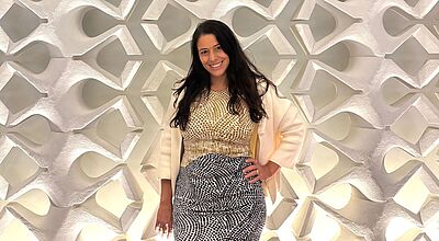 Auf Luxusunterkünfte spezialisiert: Smiling-House-Chefin Moriya Rockmann. Eine Frau steht vor einer dekorativen, beleuchteten Wand mit geometrischem Muster. Sie trägt ein gelb-weiß gemustertes Oberteil, einen cremefarbenen Blazer und einen schwarz-weiß gemusterten Rock.