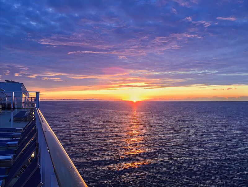 Sonnenaufgang über dem offenen Meer, aufgenommen vom Deck eines Kreuzfahrtschiffs, mit orange-violett gefärbtem Himmel und der Spiegelung der Sonne auf den Wellen.