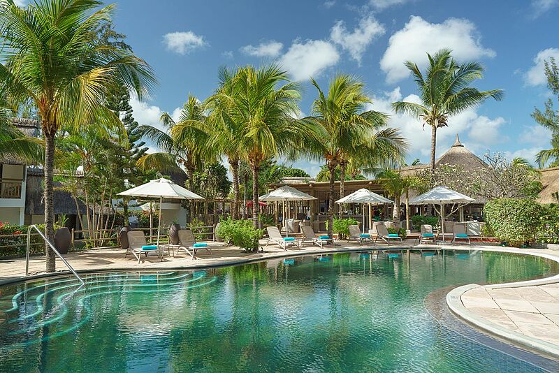 Das Coin de Mire Attitude auf Mauritius war das erste Haus der Gruppe und Vorbild für nachhaltigen, authentischen Inselurlaub. Zu sehen ist die Poolanlage eines Resorts, Liegen mit Sonnenschirmen im Vorder- Palmen und blauer Himmel im Hintergrund.
