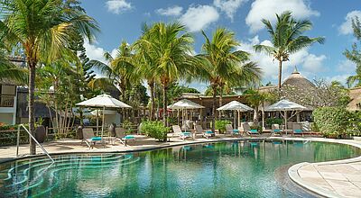 Zu sehen ist die Poolanlage eines Resorts, Liegen mit Sonnenschirmen im Vorder- Palmen und blauer Himmel im Hintergrund.