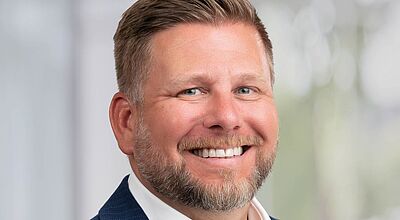 Ein lächelnder Mann mit kurzem, hellbraunem Haar und Bart trägt ein dunkelblaues Sakko über einem weißen Hemd. Der Hintergrund ist unscharf und hell, vermutlich in einem Büro oder Konferenzraum aufgenommen.