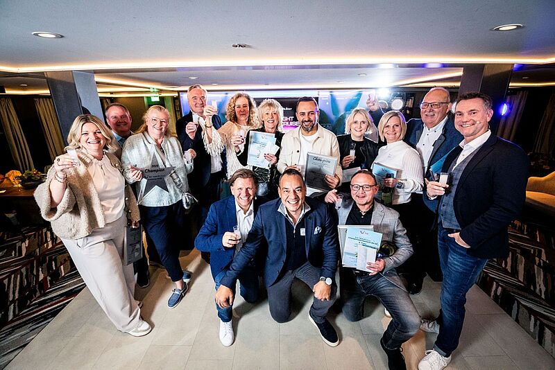 Für die „Besten der Besten“ von TUI Travel Star geht es ins Grecotel Luxme White auf Kreta, gesponsert von TUI „Gruppenfoto von etwa fünfzehn lächelnden Personen in einem hellen Innenraum. Sie halten Urkunden, Trophäen und Sektgläser in den Händen, offensichtlich anlässlich einer Preisverleihung oder Ehrung. Einige knien vorne, die meisten stehen dahinter; alle wirken fröhlich und feiern gemeinsam.“