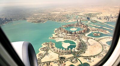 Blick aus dem Flugzeugfenster auf „The Pearl“, eine künstlich angelegte Insel in Doha, Katar. Die Lagunenartige Struktur ist von modernen Hochhäusern, Stränden und türkisfarbenem Wasser umgeben. Im Vordergrund ist das Triebwerk des Flugzeugs sichtbar.
