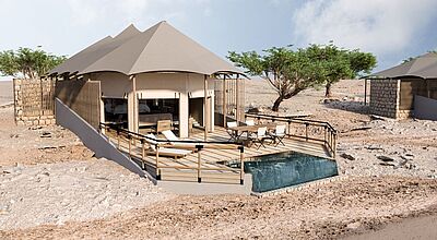Ein luxurioses Zelt, davon eine Terrasse und ein kleiner Plunge-Pool mit blauem Wasser. Ansonsten ist alles in Naturfarben. Im Hintergrund sind einige Bäume, ansonsten wächst nichts. 
