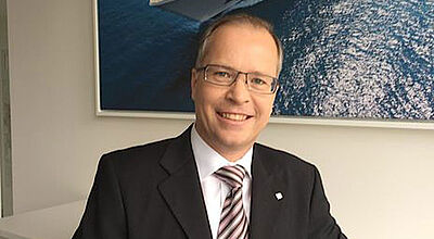 Ein Vertriebler mittleren Alters mit kurzem Haar und Brille sitzt in einem Büro. Er trägt seinen dunklen Anzug und eine rosa-braun-gestreifte Krawatte auf weißem Hemd und lächelt in die Kamera. Im Hintergrund hängt ein Bild von einem weißen Kreuzfahrtschiff. 