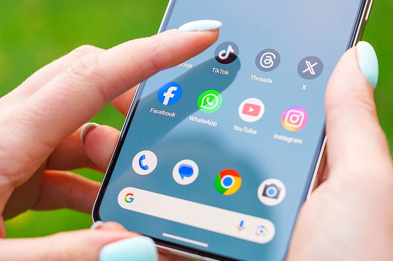 Das Bild zeigt ein Smartphone, das von einer Person mit pastellblauen Fingernägeln gehalten wird, während sie eine Social-Media-App auswählt. Sichtbar sind bekannte Apps wie TikTok, Facebook, WhatsApp, Instagram und YouTube auf dem Homescreen.