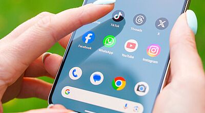 Um das Thema Social Media und Marketing dreht sich eine Infoveranstaltung von Reisezukunft. Das Bild zeigt ein Smartphone, das von einer Person mit pastellblauen Fingernägeln gehalten wird, während sie eine Social-Media-App auswählt. Sichtbar sind bekannte Apps wie TikTok, Facebook, WhatsApp, Instagram und YouTube auf dem Homescreen.