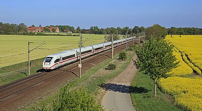 Mit dem neuen Modell entscheidet der Kunde nach der Buchung, welchen Zug er nehmen möchte. Foto: Deutsche Bahn Ein moderner ICE-Zug der Deutschen Bahn fährt an einem sonnigen Tag durch eine ländliche Landschaft mit blühenden Rapsfeldern und grünen Wiesen. Links neben der Bahnstrecke ist ein großes Gebäude mit roten Dächern zu sehen. Der Himmel ist klar und blau, während eine kleine Landstraße parallel zur Bahnstrecke verläuft.