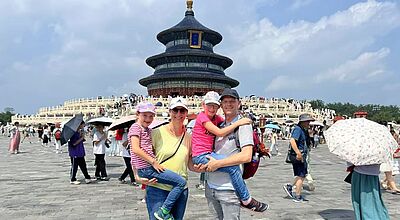 Eine Familie mit zwei Kindern posiert fröhlich vor dem Himmelstempel (Tempel des Himmels) in Peking, China. Die Eltern halten jeweils eines der Kinder auf dem Arm. Im Hintergrund sind zahlreiche Besucher sowie das markante, runde Gebäude des Himmelstempels mit seinem blauen, dreistufigen Dach zu sehen. Es ist ein sonniger Tag mit leicht bewölktem Himmel, viele Menschen schützen sich mit Schirmen vor der Sonne.