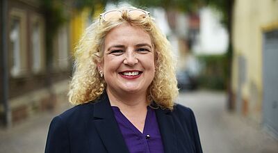 Eine Frau mit lockigem blondem Haar steht lächelnd in einer Gasse im Freien. Sie trägt ein dunkelblaues Jackett und eine violette Bluse.