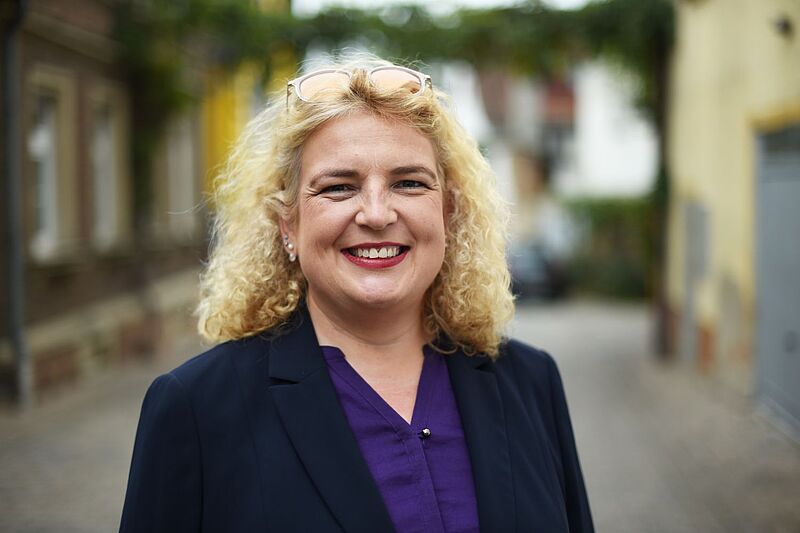 Eine Frau mit lockigem blondem Haar steht lächelnd in einer Gasse im Freien. Sie trägt ein dunkelblaues Jackett und eine violette Bluse.