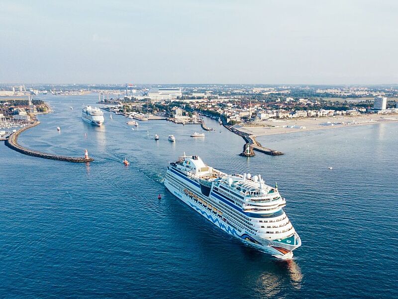 Luftaufnahme eines Kreuzfahrtschiffs von Aida Cruises beim Auslaufen in Warnemünde. Die Sonne scheint.