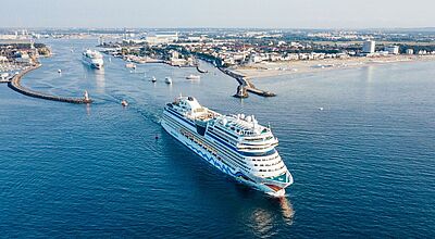 Luftaufnahme eines Kreuzfahrtschiffs von Aida Cruises beim Auslaufen in Warnemünde. Die Sonne scheint.