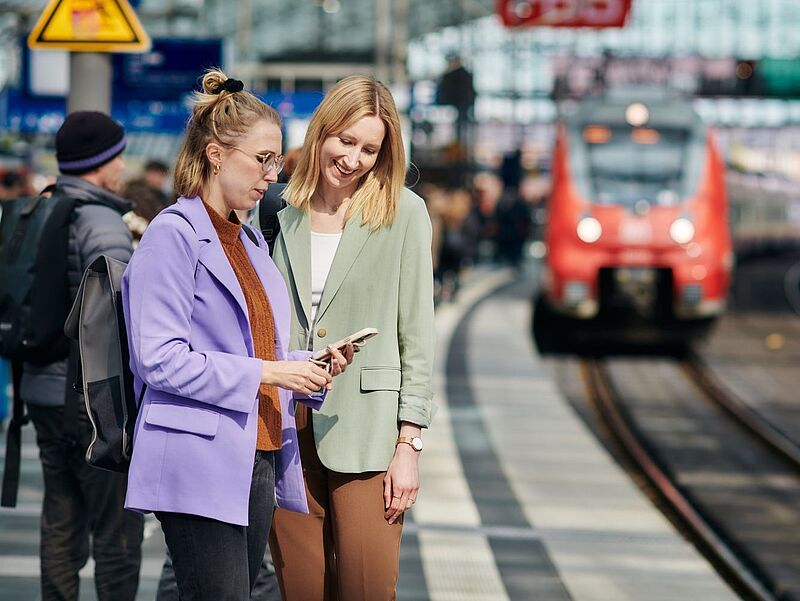 Zwei blonde Frauen stehen an einem großen Bahnhof am Bahnsteig und schauen auf ein Handy, im Hintergrund ist unscharf ein einfahrender Zug zu sehen.