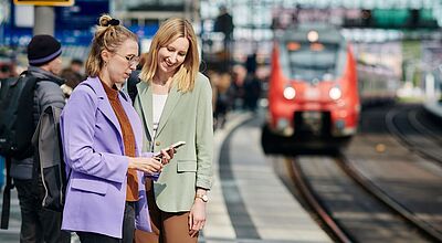 Zwei blonde Frauen stehen an einem großen Bahnhof am Bahnsteig und schauen auf ein Handy, im Hintergrund ist unscharf ein einfahrender Zug zu sehen.