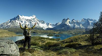 Ein Wanderer mit grünem Rucksack und gelber Jacke steht auf einem Felsen und streckt triumphierend die Arme in die Höhe. Vor ihm erstreckt sich die atemberaubende Landschaft des Torres del Paine Nationalparks in Chile mit schneebedeckten Gipfeln, tiefblauen Seen und weitläufigen Ebenen unter einem klaren, strahlend blauen Himmel. Links im Vordergrund ein großer Felsblock, rechts einige kahle Bäume.