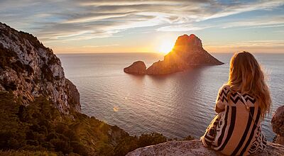 Frau sitzt auf einem Felsen und blickt bei Sonnenuntergang auf die vor der Küste liegende Felseninsel Es Vedrà vor Ibiza, Spanien.