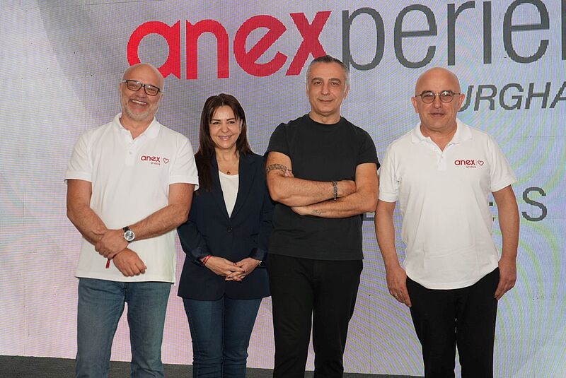 Vier Personen stehen nebeneinander vor einer großen Leinwand mit dem roten Anex-Logo. Zwei der Personen tragen weiße Polohemden mit Anex-Aufdruck, eine Person steht in dunkler Kleidung mit verschränkten Armen und eine Person trägt ein dunkelblaues Jacket. Alle schauen lächelnd in die Kamera.