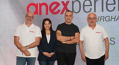 Vier Personen stehen nebeneinander vor einer großen Leinwand mit dem roten Anex-Logo. Zwei der Personen tragen weiße Polohemden mit Anex-Aufdruck, eine Person steht in dunkler Kleidung mit verschränkten Armen und eine Person trägt ein dunkelblaues Jacket. Alle schauen lächelnd in die Kamera.