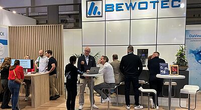 Mehrere Menschen stehen und sitzen an modernen Messeständen der Firma Bewotec auf einer Veranstaltung; es gibt Gespräche an Stehtischen und eine Theke mit Barhockern, der Firmenname „Bewotec“ ist groß auf der Wand hinter dem Stand sichtbar, einige Infomaterialien und ein Blumenarrangement sind auf den Tischen platziert.