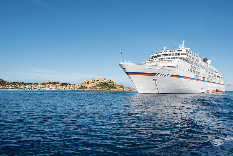 Die Reisen der Europa (Foto) sind erstmals mit denen der Europa 2 in einem Katalog zusammengefasst Das kleine Kreuzfahrtschiff „Europa“ mit weißem Anstrich und einem blau-orangenen Querstreifen über die gesamte Schiffsläge ankert bei Sonnenschein nahe einem südeuropäisch anmutenden Hafen.