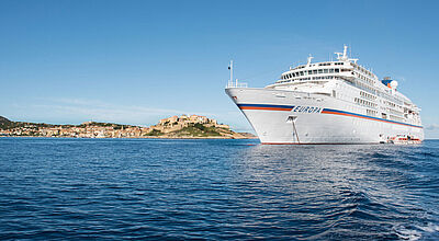 Die Reisen der Europa (Foto) sind erstmals mit denen der Europa 2 in einem Katalog zusammengefasst Das kleine Kreuzfahrtschiff „Europa“ mit weißem Anstrich und einem blau-orangenen Querstreifen über die gesamte Schiffsläge ankert bei Sonnenschein nahe einem südeuropäisch anmutenden Hafen.