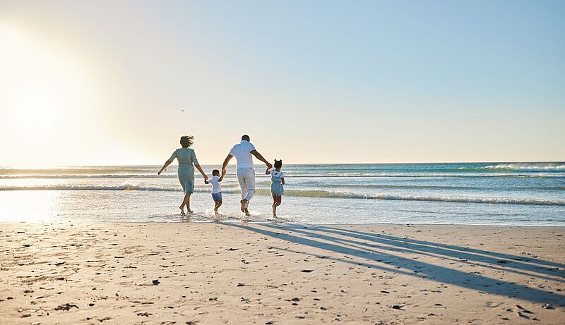 Eine von hinten fotografierte vierköpfige Familie hält sich an den Händen und läuft an einem Sandstrand in die Brandung hinein 